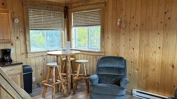L'intérieur d'un chalet en bois comprend une table à manger, des chaises, un canapé bleu, un ventilateur de plafond et une fenêtre avec des stores.