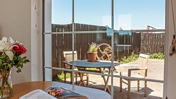 A room with a large window, a wooden table with a book, a vase with flowers, and a view of a wooden fence and outdoor furniture. - Higher Stancombe Holiday Cottages (Kingsbridge)