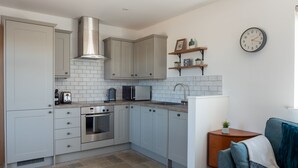 A compact kitchen with grey cabinets, a stainless steel range hood, a built-in oven, and a microwave. There is a wooden sideboard with a plant, and a clock on the wall. - Higher Stancombe Holiday Cottages (Kingsbridge)