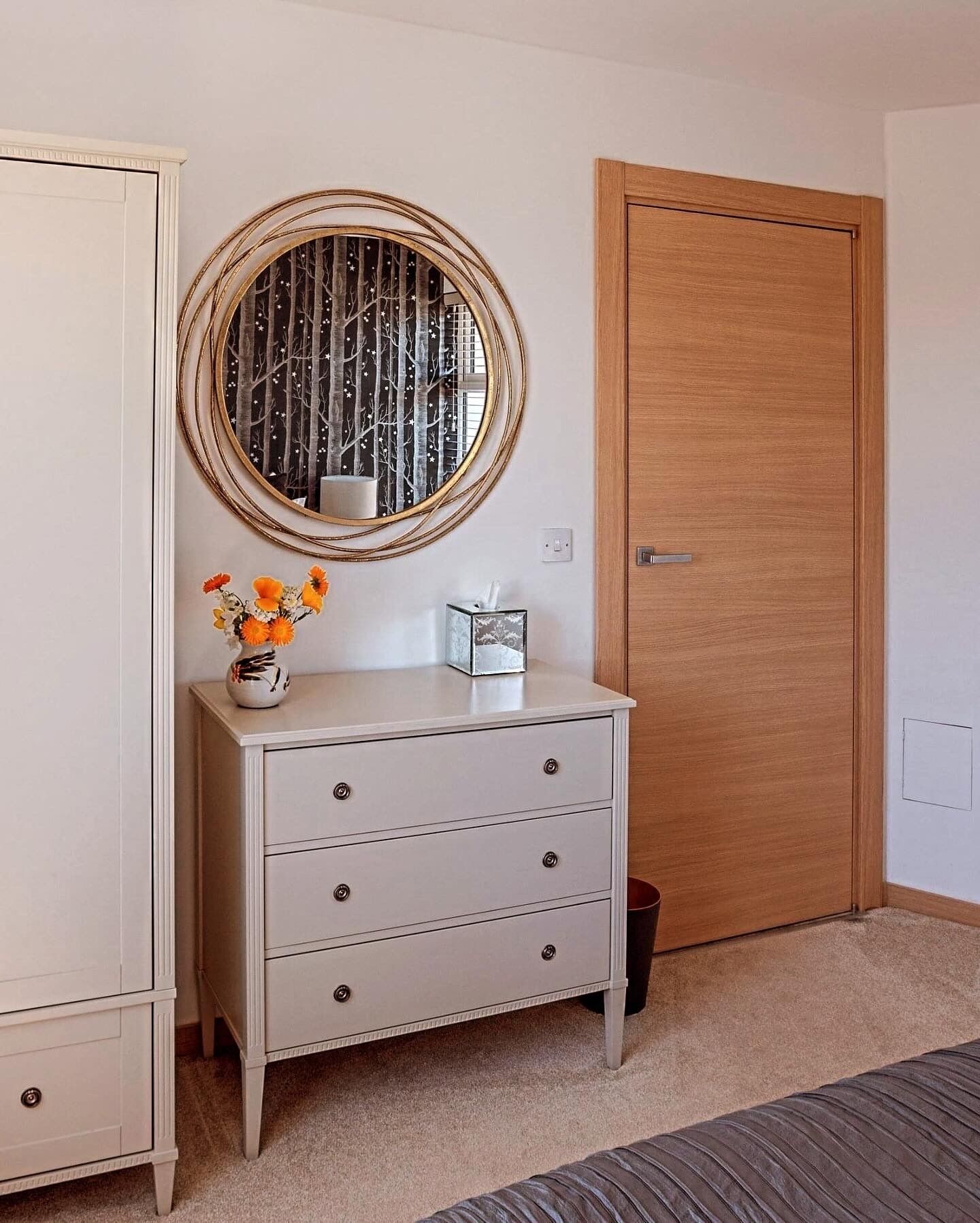 A room with a white dresser, a round mirror with a decorative frame, a vase with flowers, and a wooden door.