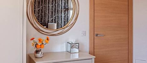 A room with a white dresser, a round mirror with a decorative frame, a vase with flowers, and a wooden door.