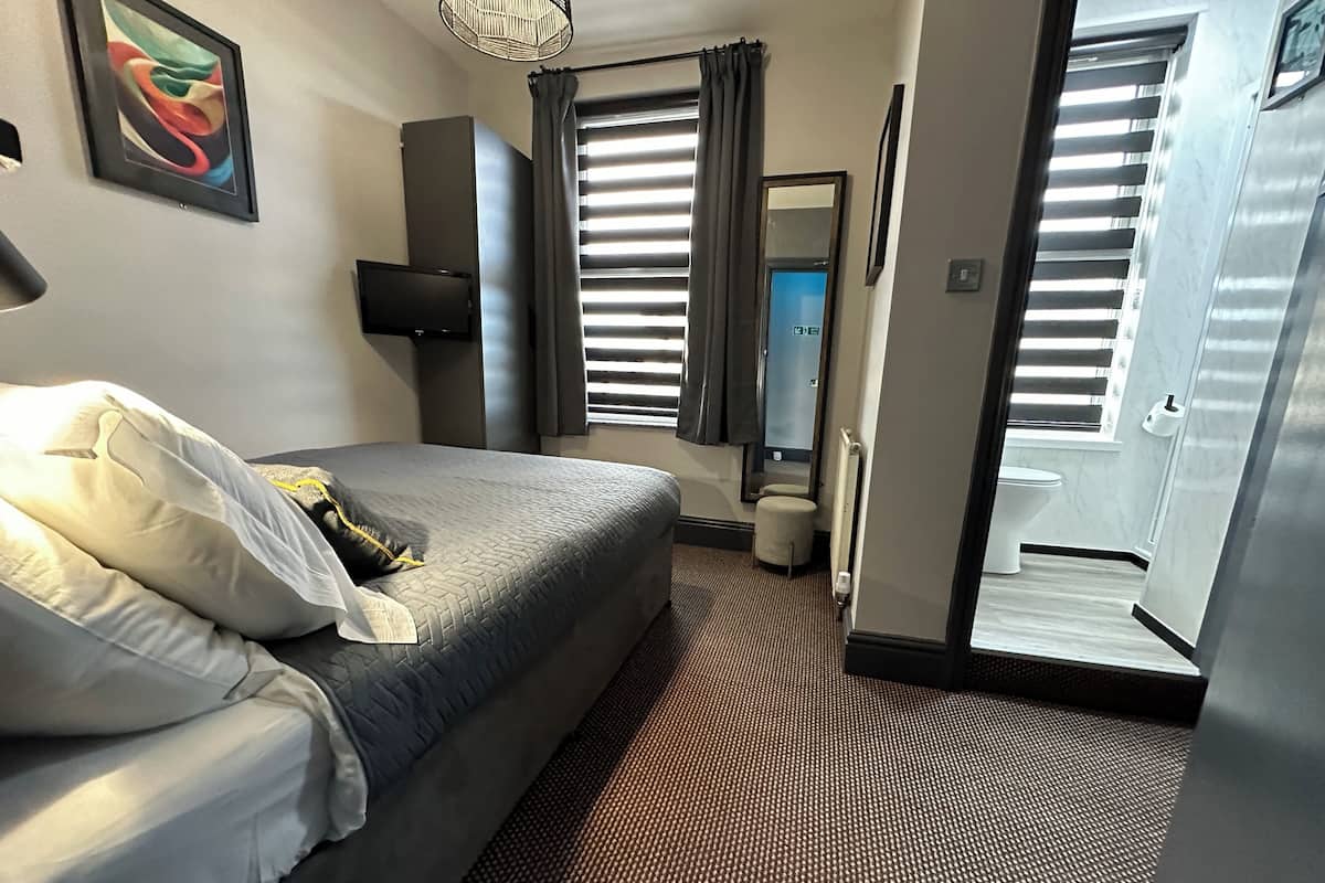 A hotel room with a bed, a bedside table, a lamp, a window with blinds, and a door leading to another room.