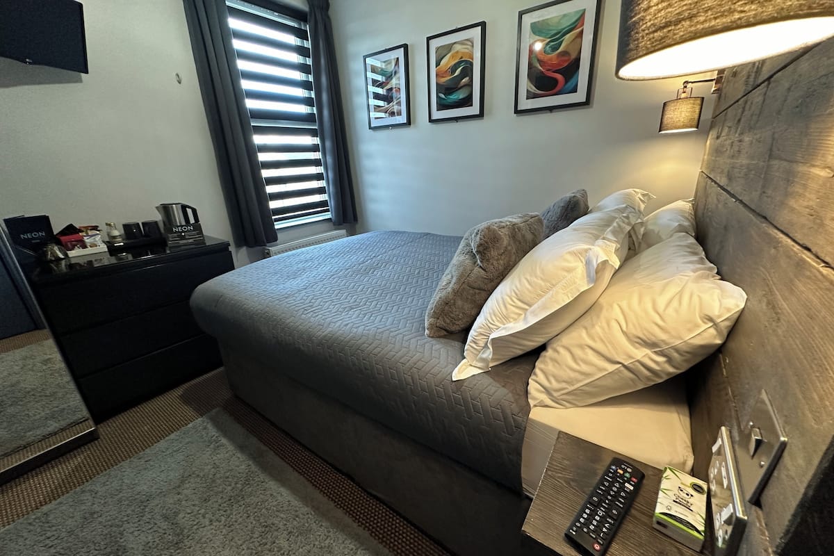A neatly arranged bedroom with a bed, pillows, a bedside table with a remote control, a lamp, and artwork on the wall.