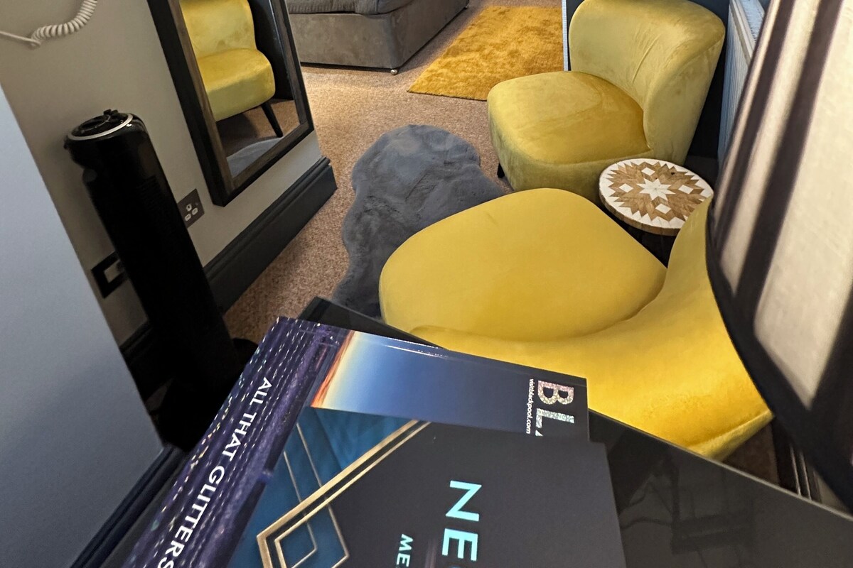A compact hotel room with a bed, a seating area with a yellow chair and ottoman, a small round table, a mirror, and a ceiling light.