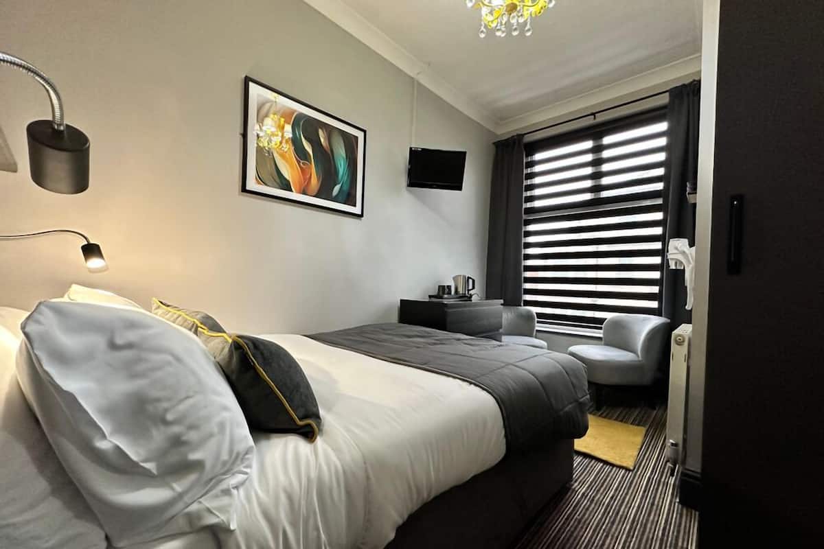 A modern hotel room with a large bed, a desk, a chair, and a window with blinds.