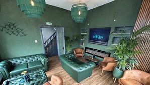 A modern lounge area with green leather sofas, a coffee table, and brown leather chairs. A flat-screen TV is mounted on the wall, and there are potted plants around. - Hotel Neon (Blackpool)