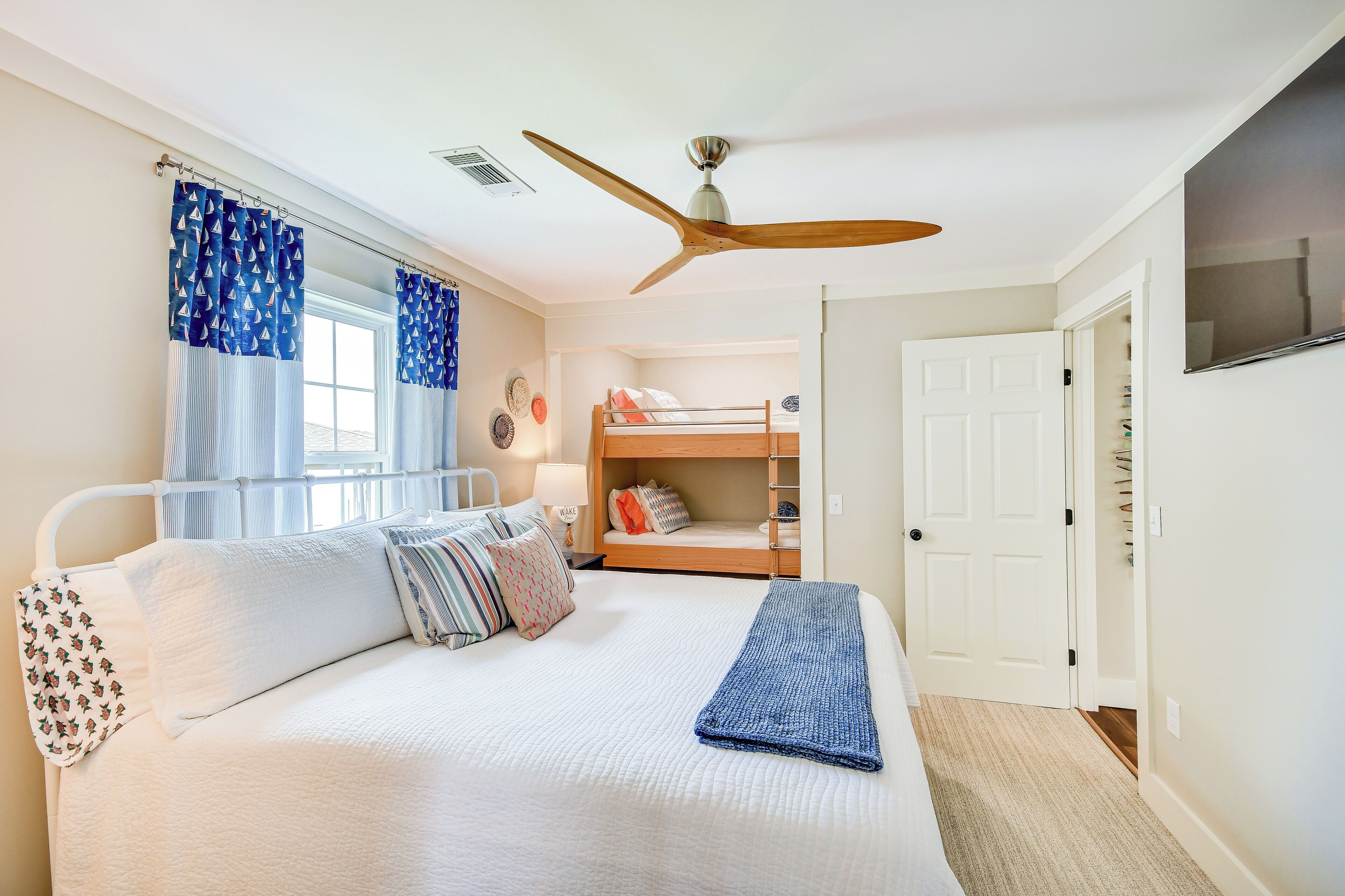 Une chambre avec un lit superposé, un ventilateur de plafond, un lit avec une literie blanche et des oreillers à motifs, et une fenêtre avec des rideaux bleus.