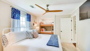 A bedroom with a bunk bed, a ceiling fan, a bed with white bedding and patterned pillows, and a window with blue curtains.