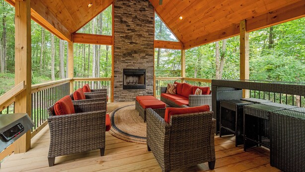 A covered outdoor area with wicker furniture, a stone fireplace, and a view of the forest. - Black Diamond Lodge by Taylor Made Deep Creek (McHenry)