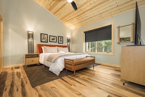 A bedroom with a wooden ceiling, a bed with white bedding, a nightstand, a dresser, and a window with a view of trees. - Black Diamond Lodge by Taylor Made Deep Creek (McHenry)
