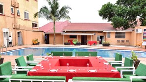 A poolside dining area with green chairs and a red tablecloth, surrounded by a pool and a building with a red roof. - Admiral hotel (Kampala)