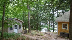 A cabin with a small porch and a window, surrounded by dense forest and a body of water. - Muskoka Serenity - Four Bedroom Cottage, Sleeps 8 (Torrance)