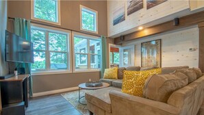 A cozy living room with a beige sofa, a coffee table, and a flat-screen TV. Large windows provide natural light and views of the outdoors. - Muskoka Serenity - Four Bedroom Cottage, Sleeps 8 (Torrance)