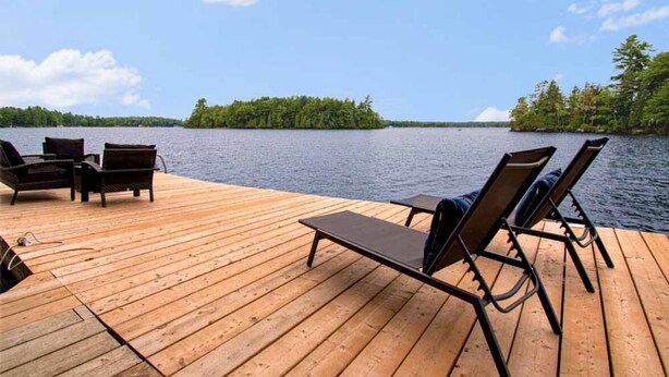 A wooden deck with lounge chairs and a table overlooking a lake with a forested island in the distance. - Muskoka Serenity - Four Bedroom Cottage, Sleeps 8 (Torrance)
