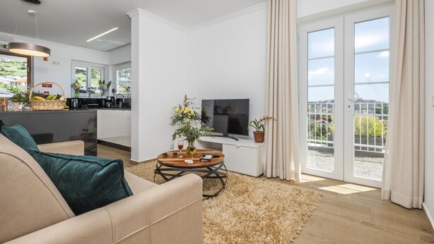 A modern living room with a sofa, a coffee table, and a TV. There is a kitchen area with a sink and a window. The room has large sliding glass doors leading to a balcony with a view of the city. - Casas da Luz - Casa da Pedreira by Homie (Ponta do Sol)