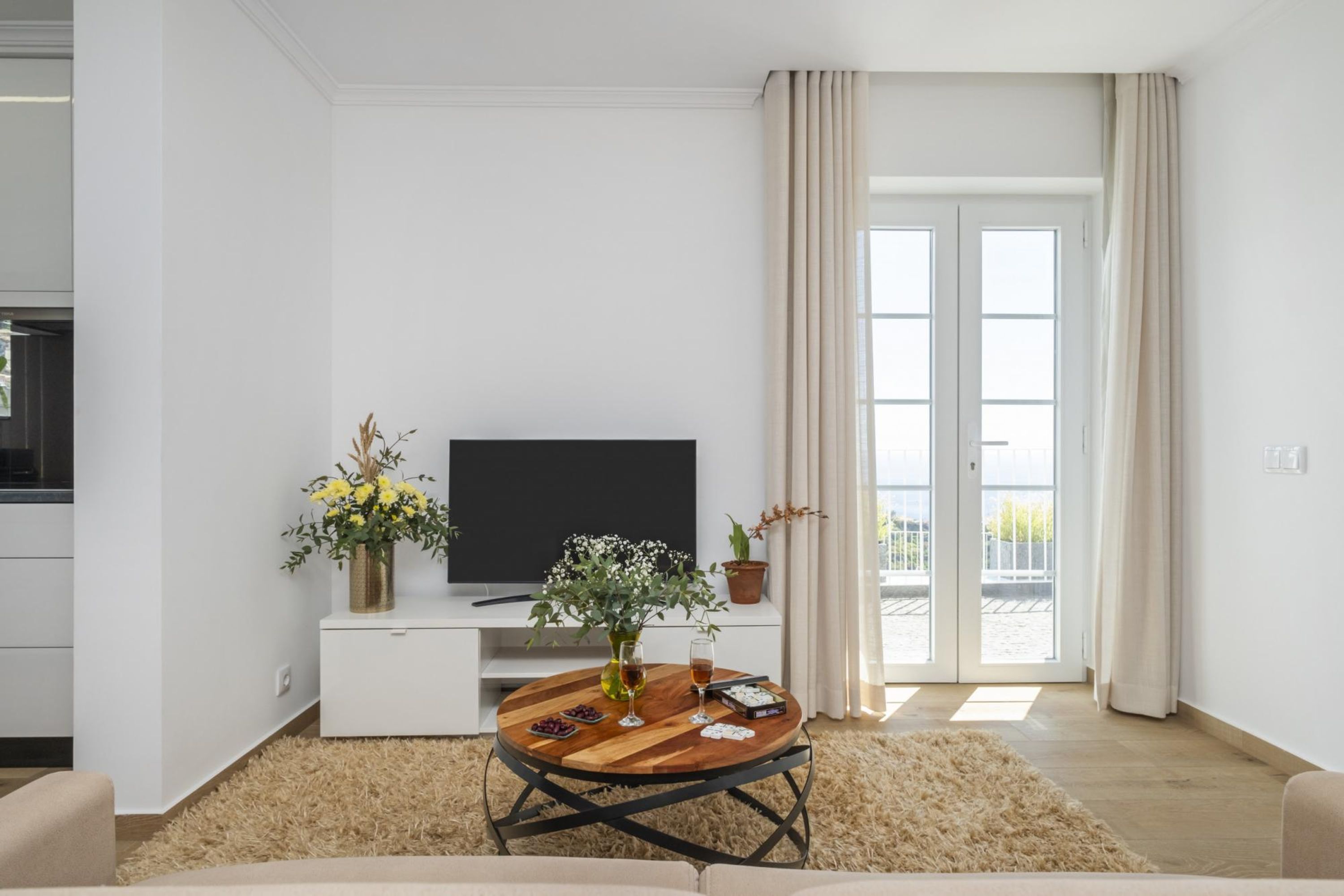Un salon moderne avec un téléviseur à écran plat, une table basse en bois et un canapé. On y trouve une grande fenêtre avec des rideaux, une plante en pot et une petite table avec des boissons et des en-cas.