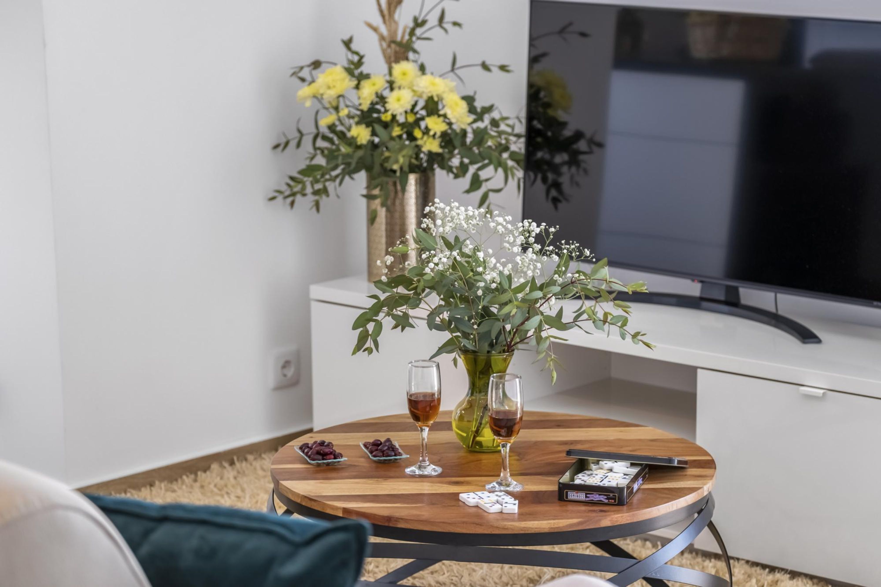 A round wooden table with two glasses of amber liquid, a deck of cards, and a small bowl of grapes. A vase with yellow flowers and white baby's breath is on the table. A flat-screen TV is mounted on the wall behind the table.