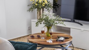 A round wooden table with two glasses of amber liquid, a deck of cards, and a small bowl of grapes. A vase with yellow flowers and white baby's breath is on the table. A flat-screen TV is mounted on the wall behind the table. - Casas da Luz - Casa da Pedreira by Homie (Ponta do Sol)