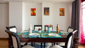 A dining area with a table set for four, surrounded by chairs, with artwork on the walls and a window with curtains. - Skyline Apartment 3 Bedroom with SQ-801 (Nairobi)