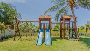 Una estructura de juego de madera con toboganes y columpios, rodeada de césped y palmeras.
