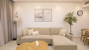 Ein modernes Wohnzimmer mit einem beigefarbenen Ecksofa, einem runden Couchtisch mit Kerzen, einer Stehlampe, einem Blumentopf und zwei gerahmten Postern von Stadtansichten an der Wand.