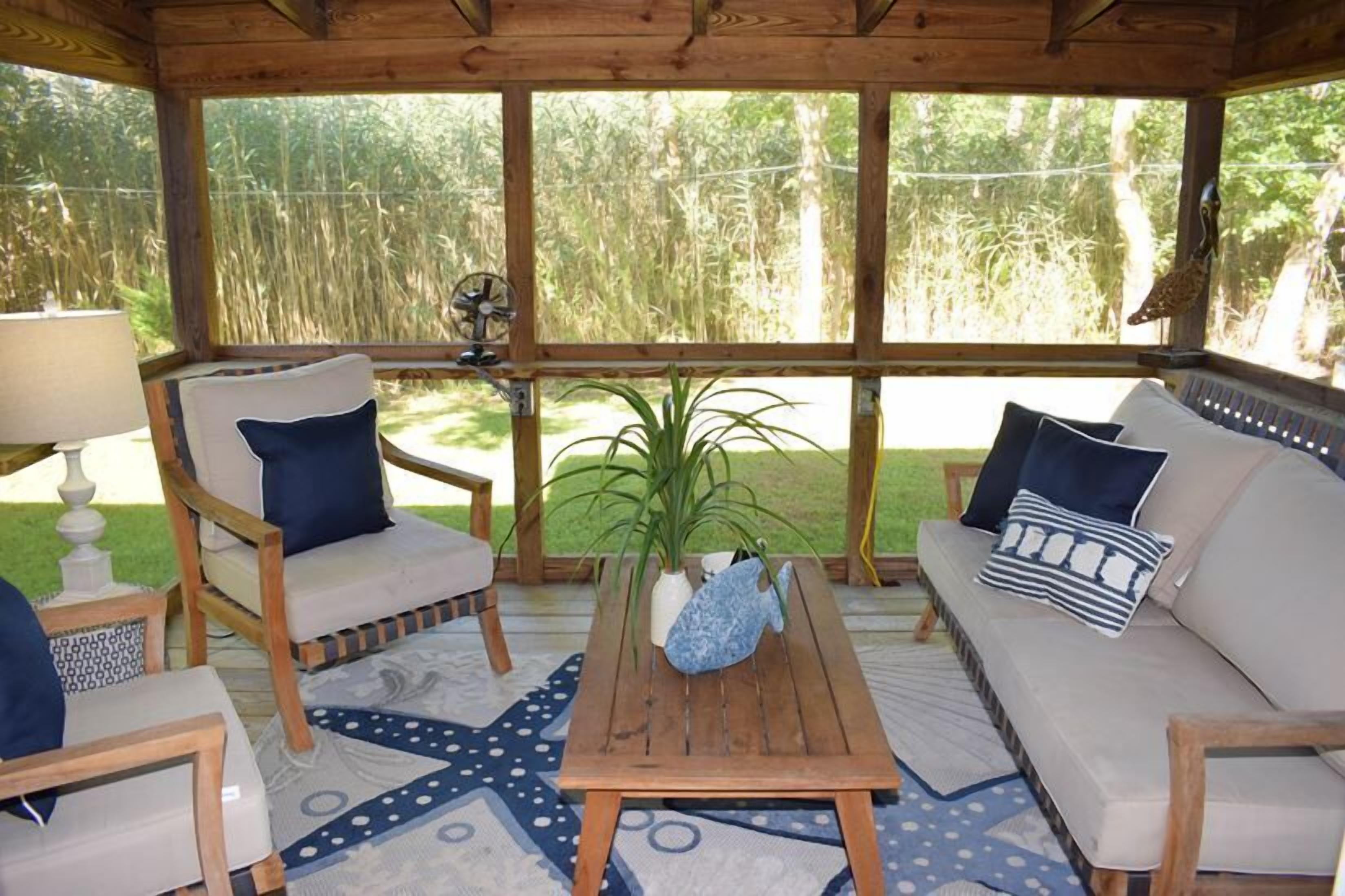 A covered outdoor seating area with a sofa, chairs, and a table, surrounded by large windows with a view of a grassy area.