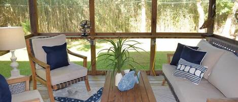 A covered outdoor seating area with a sofa, chairs, and a table, surrounded by large windows with a view of a grassy area.