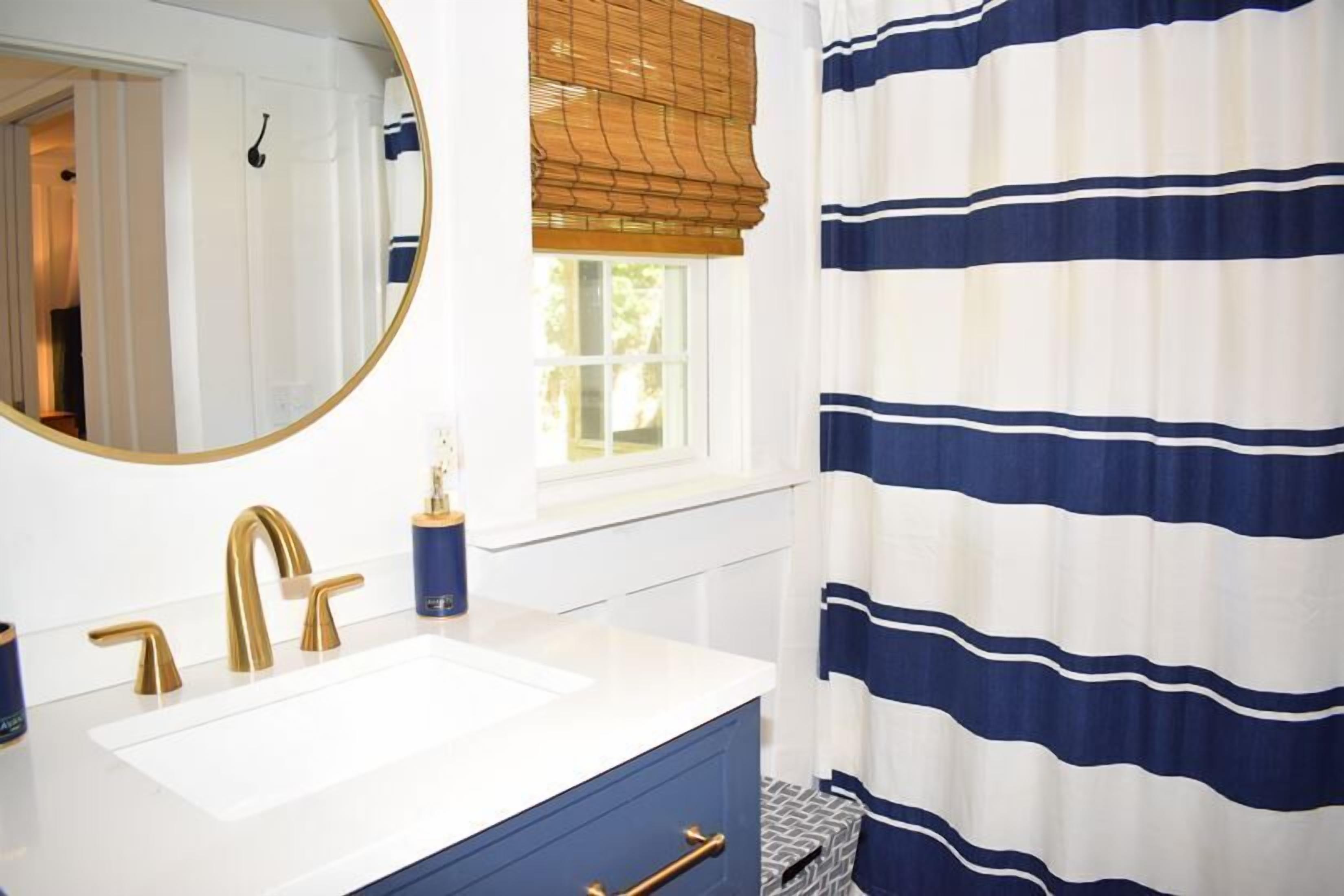 A bathroom with a white sink, gold faucets, a blue cabinet, a round mirror, and a striped shower curtain.