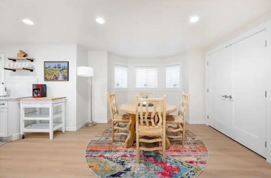 Uma área de refeições com uma mesa redonda e cadeiras, um tapete colorido e um armário branco com uma prateleira acima.