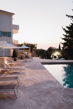 A poolside area with lounge chairs and umbrellas, a stone wall, and a building with a balcony. - Locanda Villa Lefkada A (Lefkada)
