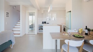 A modern kitchen with white cabinetry, a central island, and a dining area with a wooden table and white chairs. - Locanda Villa Lefkada C (Lefkada)