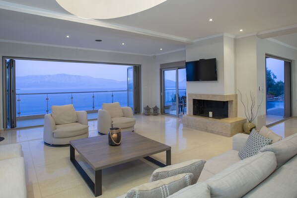 A spacious living room with a fireplace, comfortable seating, and large windows offering a view of the sea. - Villa Lilaea Lefkada (Lefkada)