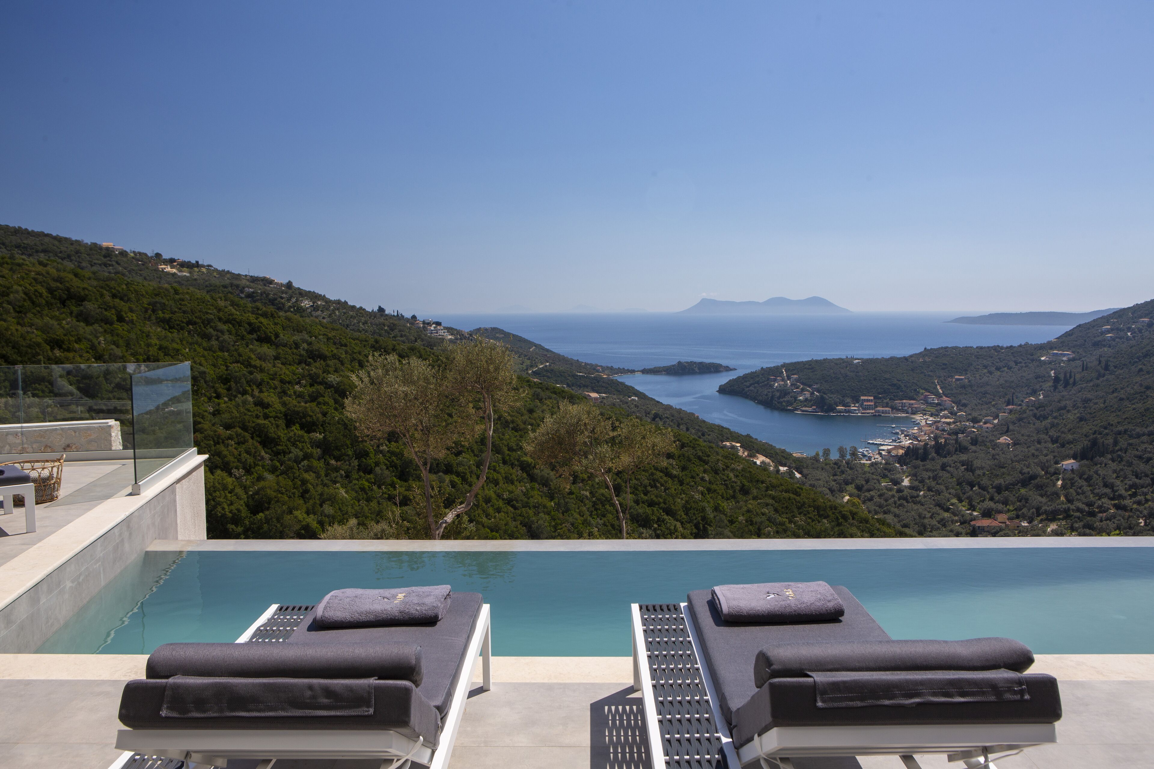 An infinity pool with two lounge chairs, overlooking a coastal landscape with hills and a clear view of the sea.