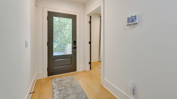 A hallway with a wooden floor, a gray rug, a dark door with a glass panel, and a white wall with a thermostat.