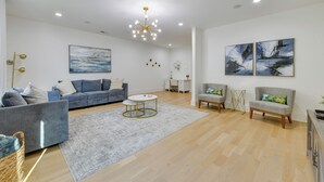 A modern living room with a grey sofa, a round coffee table, and two armchairs. There is a painting on the wall and a chandelier hanging from the ceiling.