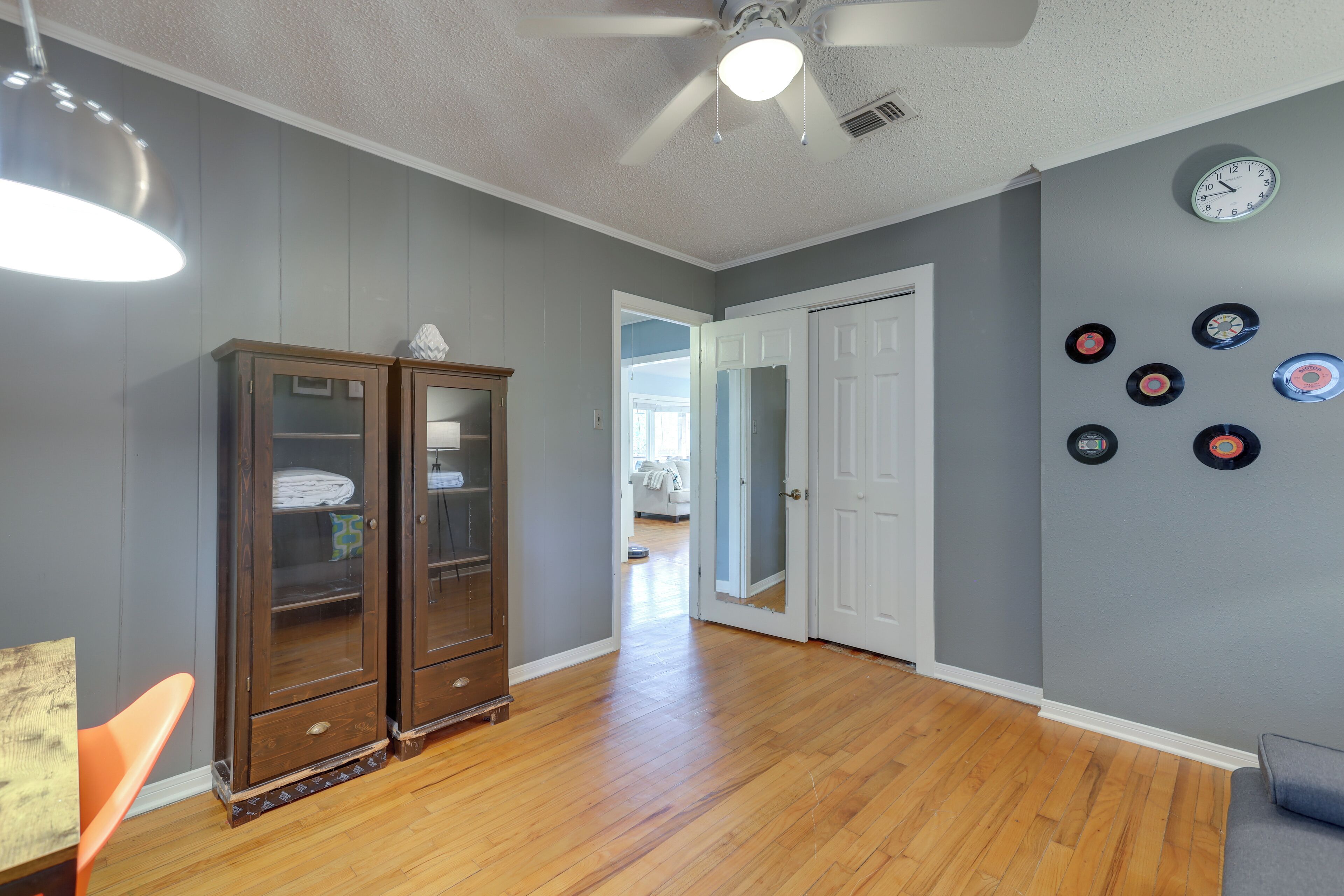 A room with wooden flooring, a ceiling fan, a cabinet with glass doors, a door, and wall-mounted records.