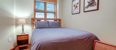 A bedroom with a bed, a nightstand, a lamp, a window with blinds, and two framed pictures on the wall.