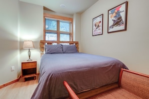 Un dormitorio con cama, mesita de noche, lámpara, ventana con persianas y dos cuadros enmarcados en la pared.