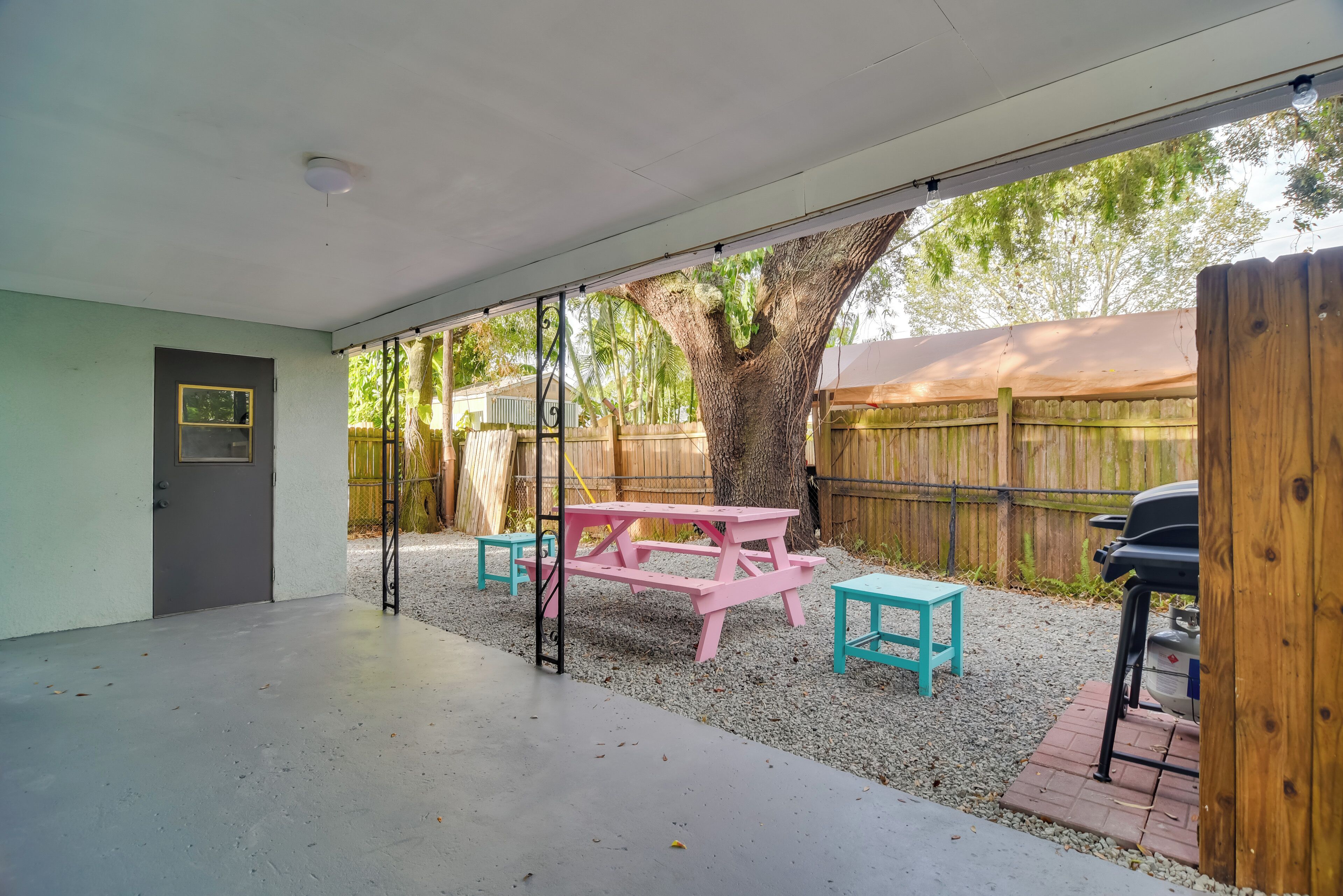 Un patio techado con mesa de picnic y bancos, un árbol y una valla de madera al fondo.