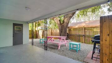 Un patio techado con mesa de picnic y bancos, un árbol y una valla de madera al fondo.