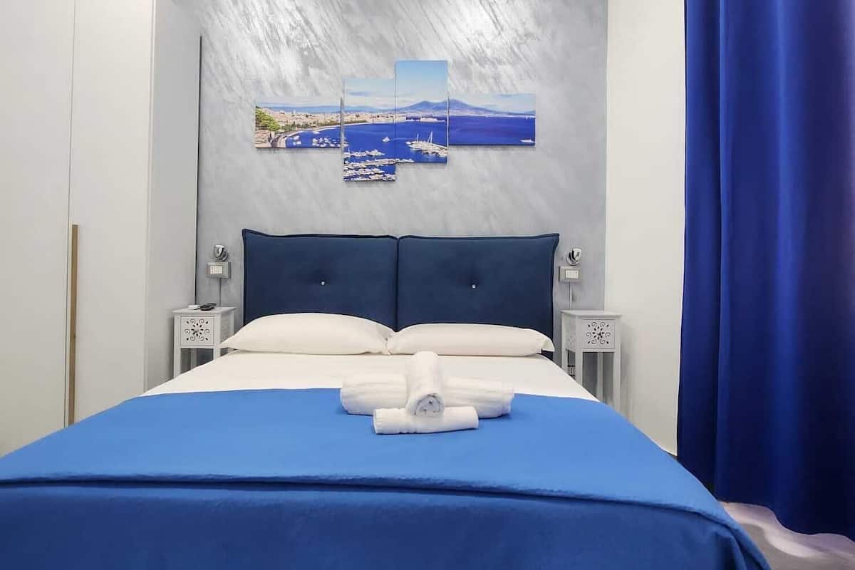 Una habitación de hotel con cama, cabecera, obra de arte colgada en la pared, mesita de noche, armario y cortina.