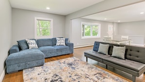 A spacious living room with a grey sofa, a dark grey sofa, and a patterned rug. There are two windows providing natural light. - Hampton Bays Retreat: Walk to Squires Beach! (Hampton Bays)