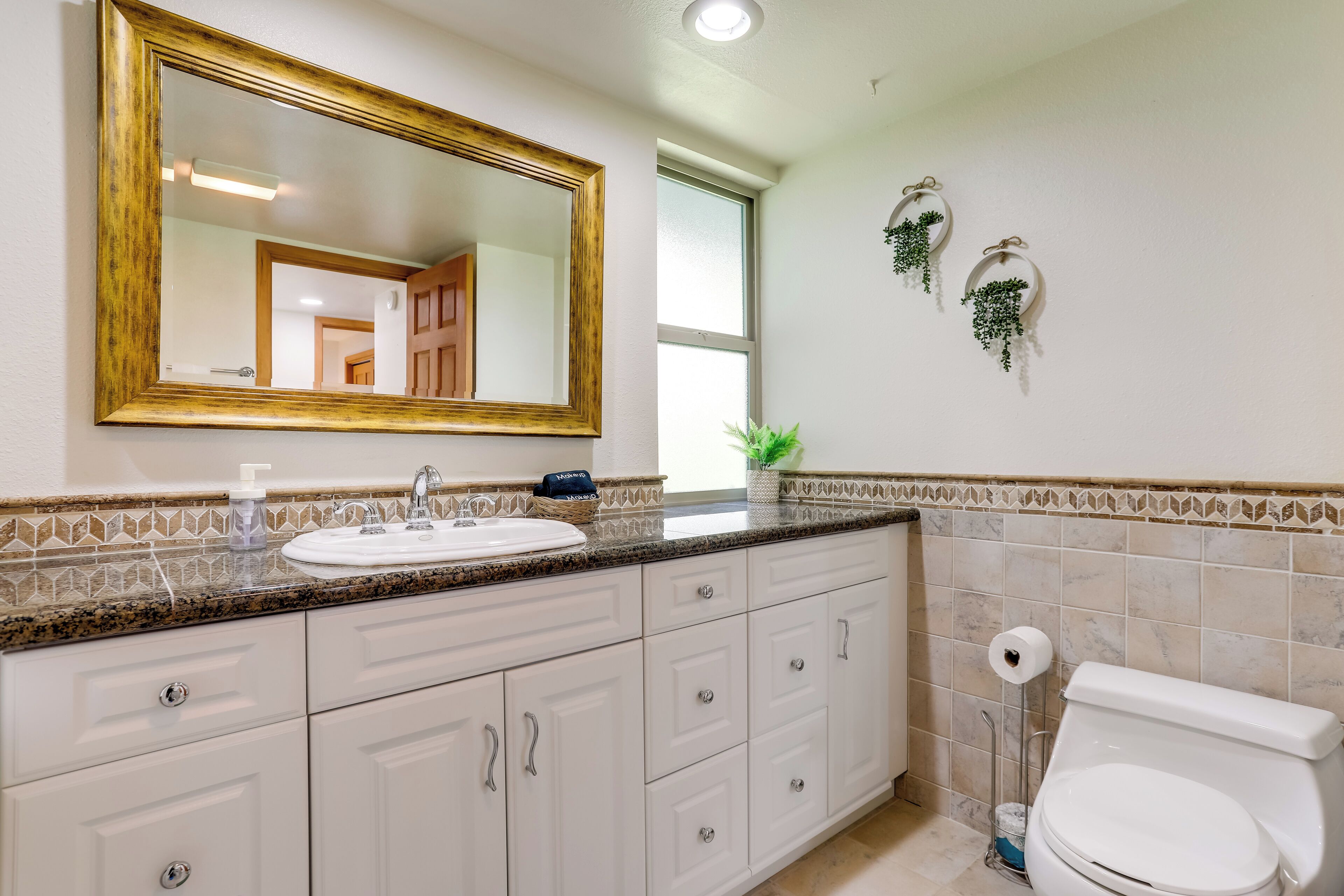 A bathroom with a large mirror, white cabinets, a sink with a soap dispenser, and a toilet.