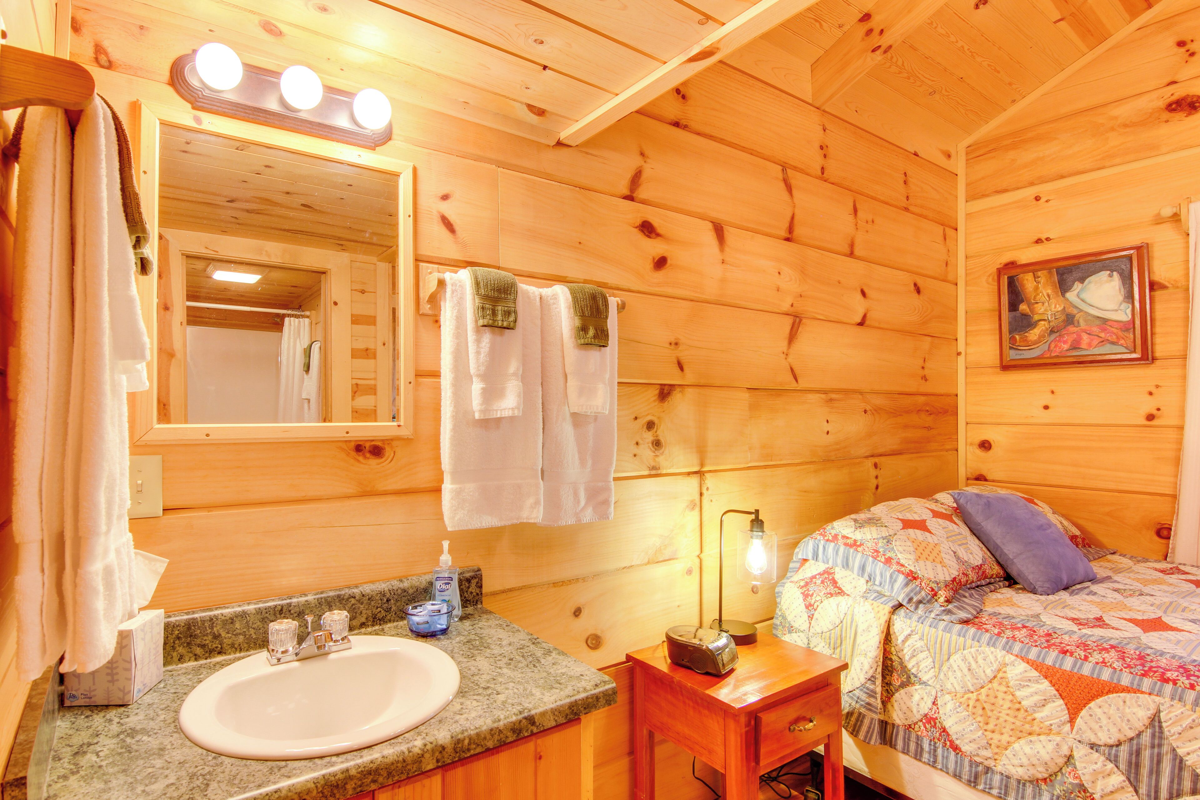 A wooden cabin room with a wooden ceiling, a sink, a mirror, a bed with a quilt, and a painting on the wall.