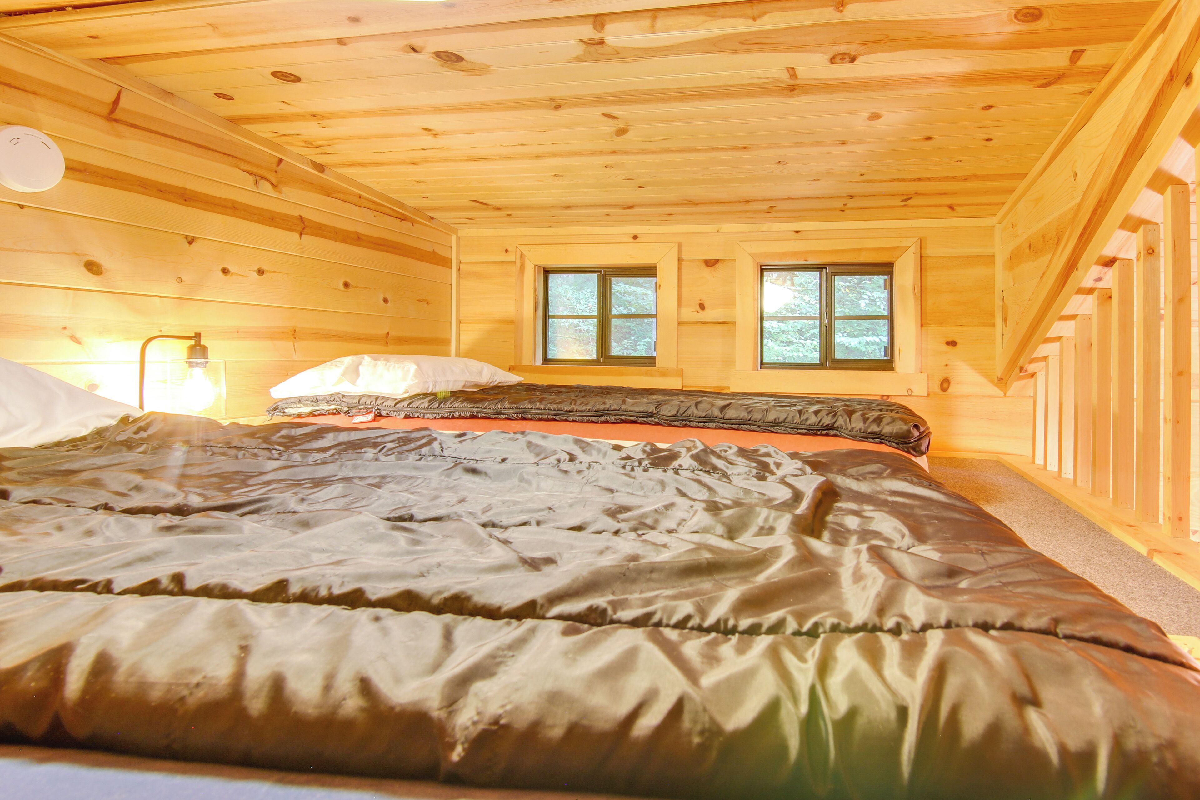 A wooden cabin bedroom with a bed, two windows, and a nightstand with a lamp.