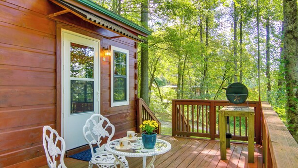 A wooden cabin with a deck, two white chairs, a small table with a vase of yellow flowers, and a view of a forest. - Hendersonville Cabin Near Hiking & Waterfalls! (Hendersonville)