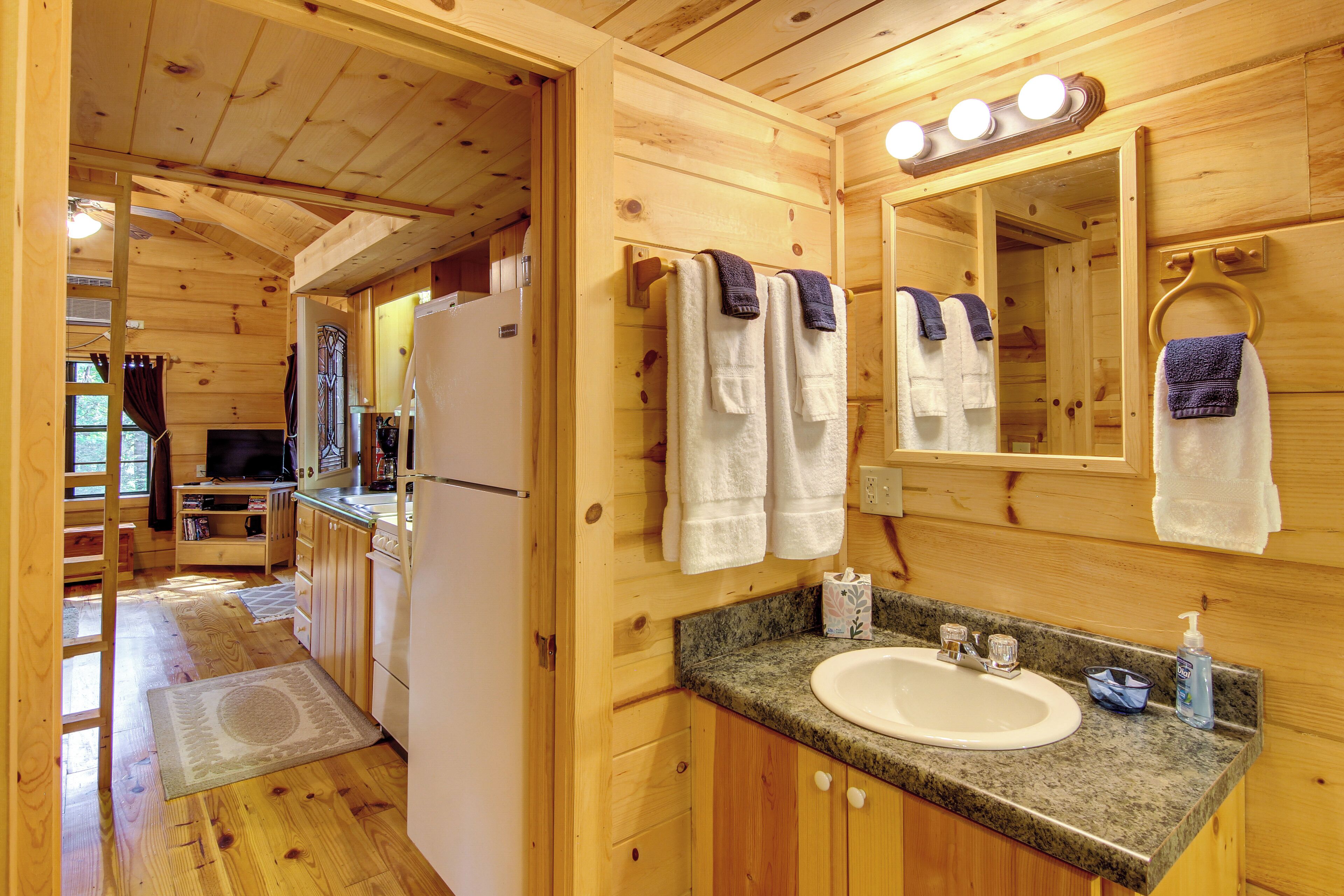 A wooden cabin interior with a kitchen, a bathroom with a sink and mirror, and a hallway leading to a bedroom.