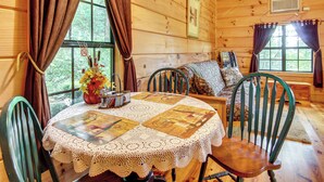 A wooden cabin interior with a round table, chairs, a quilted tablecloth, and a window with curtains. - Hendersonville Cabin Near Hiking & Waterfalls! (Hendersonville)