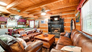 A cozy cabin interior with leather sofas, a fireplace, wooden walls, and a kitchen area.
