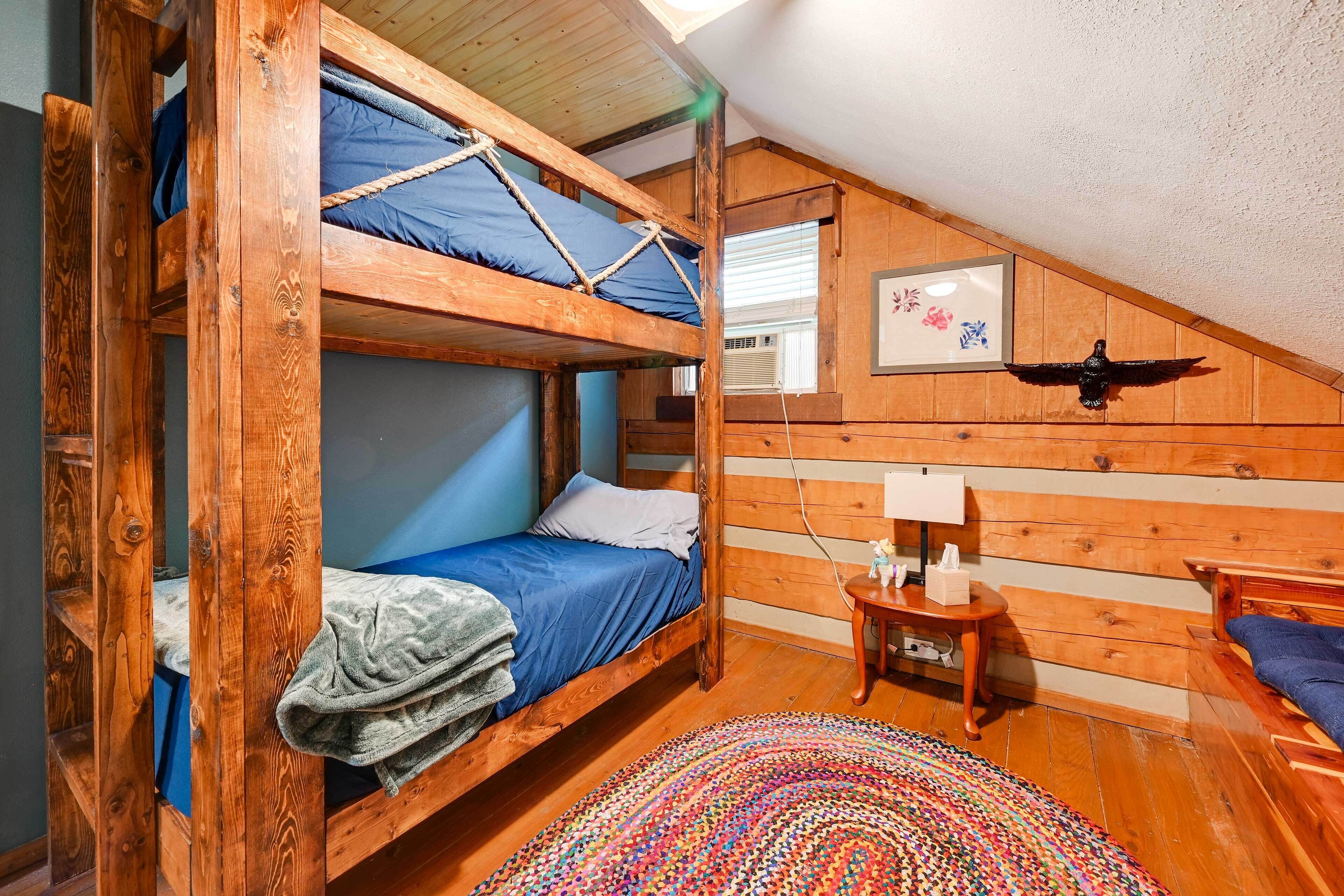 A cozy attic bedroom with wooden bunk beds, a small bedside table with a lamp, and a colorful woven rug.
